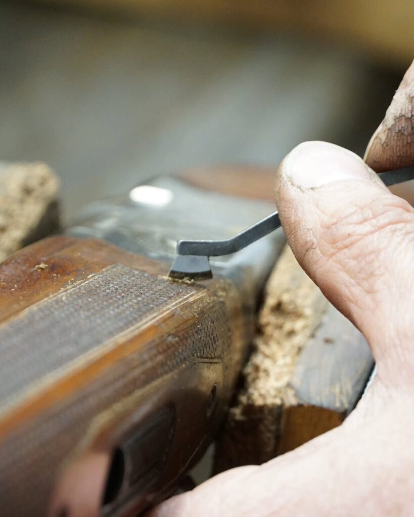 Main utilisant un outil de précision pour restaurer le quadrillage sur la crosse en bois d'une arme, avec copeaux visibles.