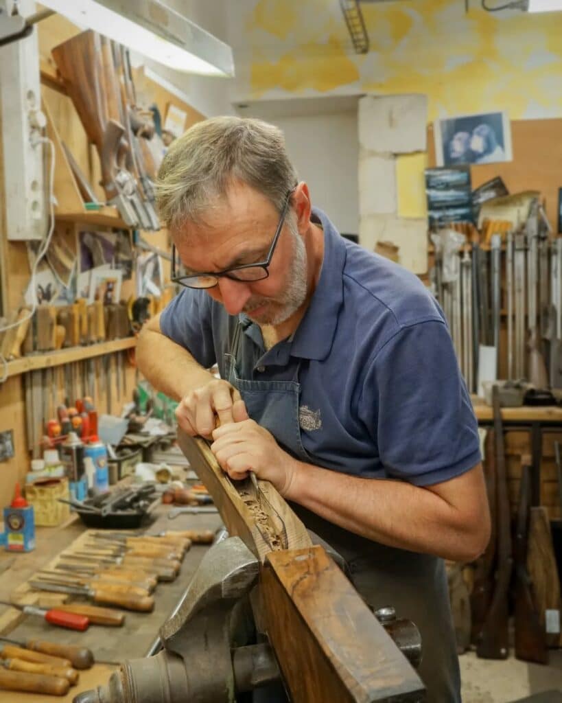 Armurier : sculpture bois d'une crosse de fusil Armurier en lunettes sculptant une crosse de fusil en bois avec un ciseau dans son atelier. Outils visibles.