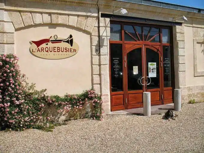 Facade avec le panneau "L'ARQUEBUSIER TONY GICQUEL" montrant une arme stilisée. Roses roses et un chien couché sur le gravier devant les portes en bois.