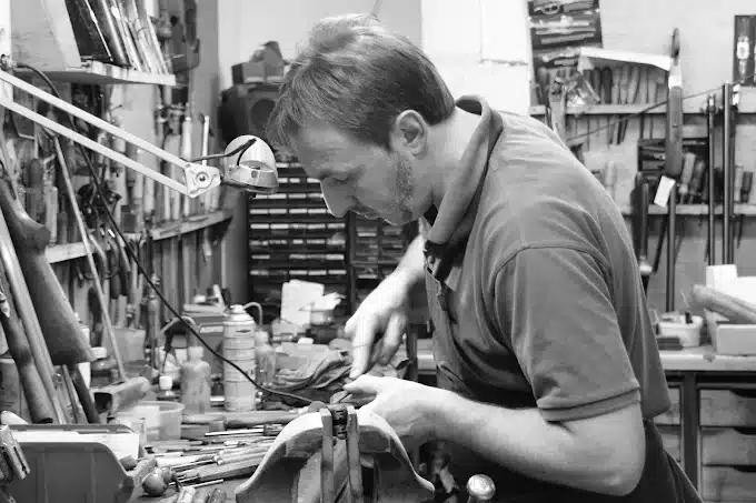 Artisan concentré travaille sur une pièce dans un étau. Nombreux outils et crosses d'armes visibles dans son atelier.