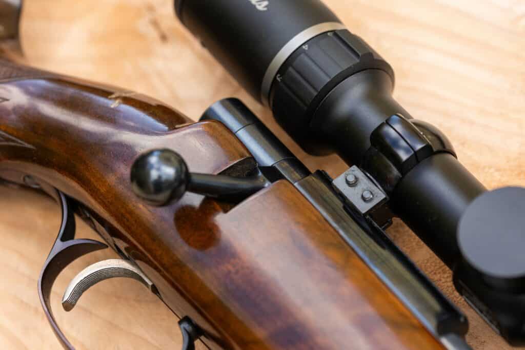 Détails d'une carabine : crosse en bois verni, culasse, détente et lunette noire avec index 3, 4. Sur fond bois.