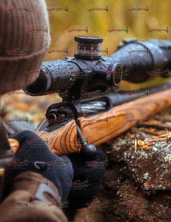 Personne aimant un fusil avec lunette sous la pluie en forêt. Gants sur la crosse bois. Filigrane L'ARQUEBUSIER visible.