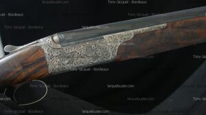 Fusil de chasse gravé : scène de gibier, volutes, crosse en bois veiné. Texte 'Tony Gicquel - Bordeaux', 'larquebusier.com'.
