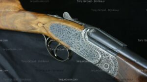 Fusil de chasse haut de gamme avec gravures florales complexes sur le boîtier métallique argenté et crosse en bois veiné. Texte : Tony Gicquel - Bordeaux.