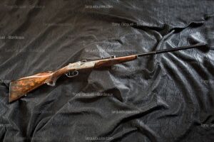Fusil de chasse ou de tir luxueux, crosse en bois veiné, canon noir long, platine gravée finement, posé sur tissu sombre froissé.