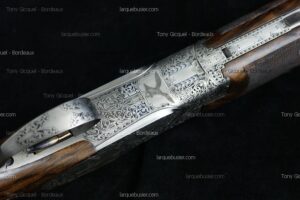 Détail d'un récepteur de fusil finement gravé avec motifs de volutes et un oiseau en vol. Fût en bois visible. Texte: Tony Gicquel - Bordeaux, larquebusier.com.