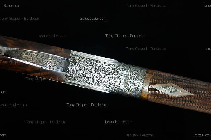 Gros plan d'une bascule de fusil de chasse à la gravure ornementale argentée et d'un fût en bois de noyer finement quadrillé.