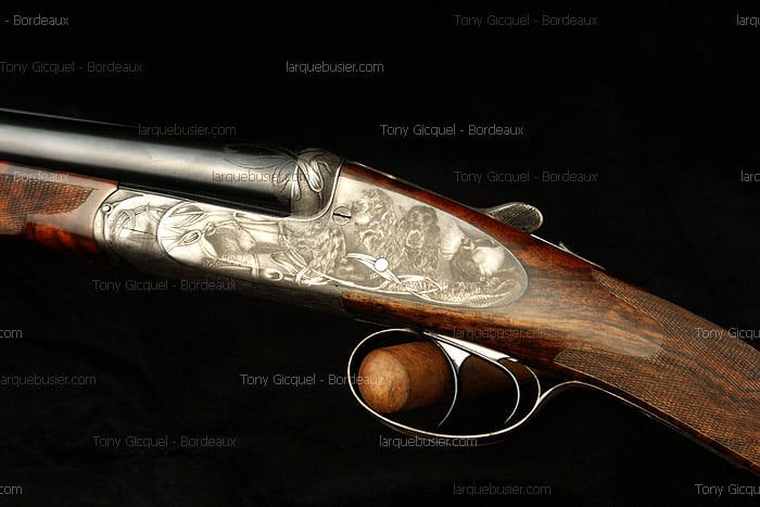 Fusil de chasse haut de gamme avec gravure détaillée sur le boîtier : trois chiens et oiseaux, crosse bois quadrillée.