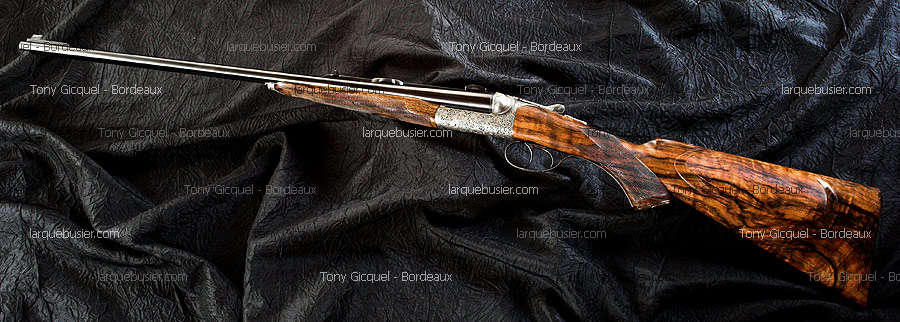 Fusil de chasse juxtaposé élégant, crosse en bois veiné et gravures argentées. Sur fond sombre. Watermarks Tony Gicquel - Bordeaux et larquebusier.com.