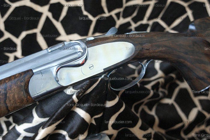 Mécanisme poli d'un fusil de chasse en bois sombre, gâchette visible, sur fond motif zèbre. Tony Gicquel - Bordeaux, larquebusier.com.