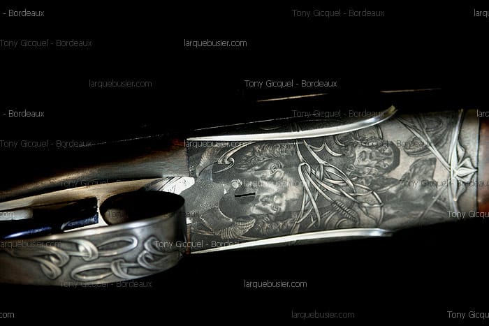 Gros plan d'un fusil orné, son récepteur en métal gravé d'un visage expressif et de motifs complexes. Mentions: Tony Gicquel - Bordeaux, larquebusier.com.