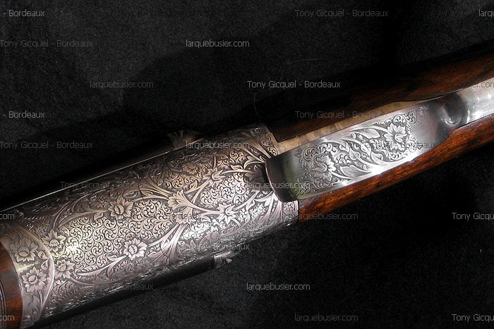 Gros plan sur la gravure florale argentée d'un fusil de chasse, avec des détails en bois sombre. Marqueurs Tony Gicquel - Bordeaux, larquebusier.com visibles.