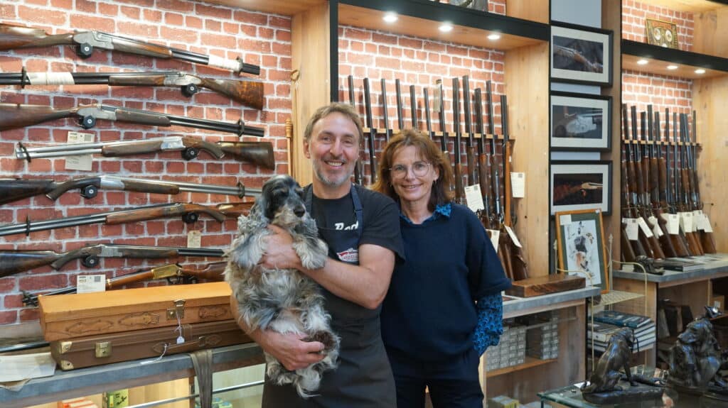 Un couple souriant avec leur chien dans une armurerie L'Arquebuserie. Nombreux fusils de chasse exposés sur un mur de briques.
