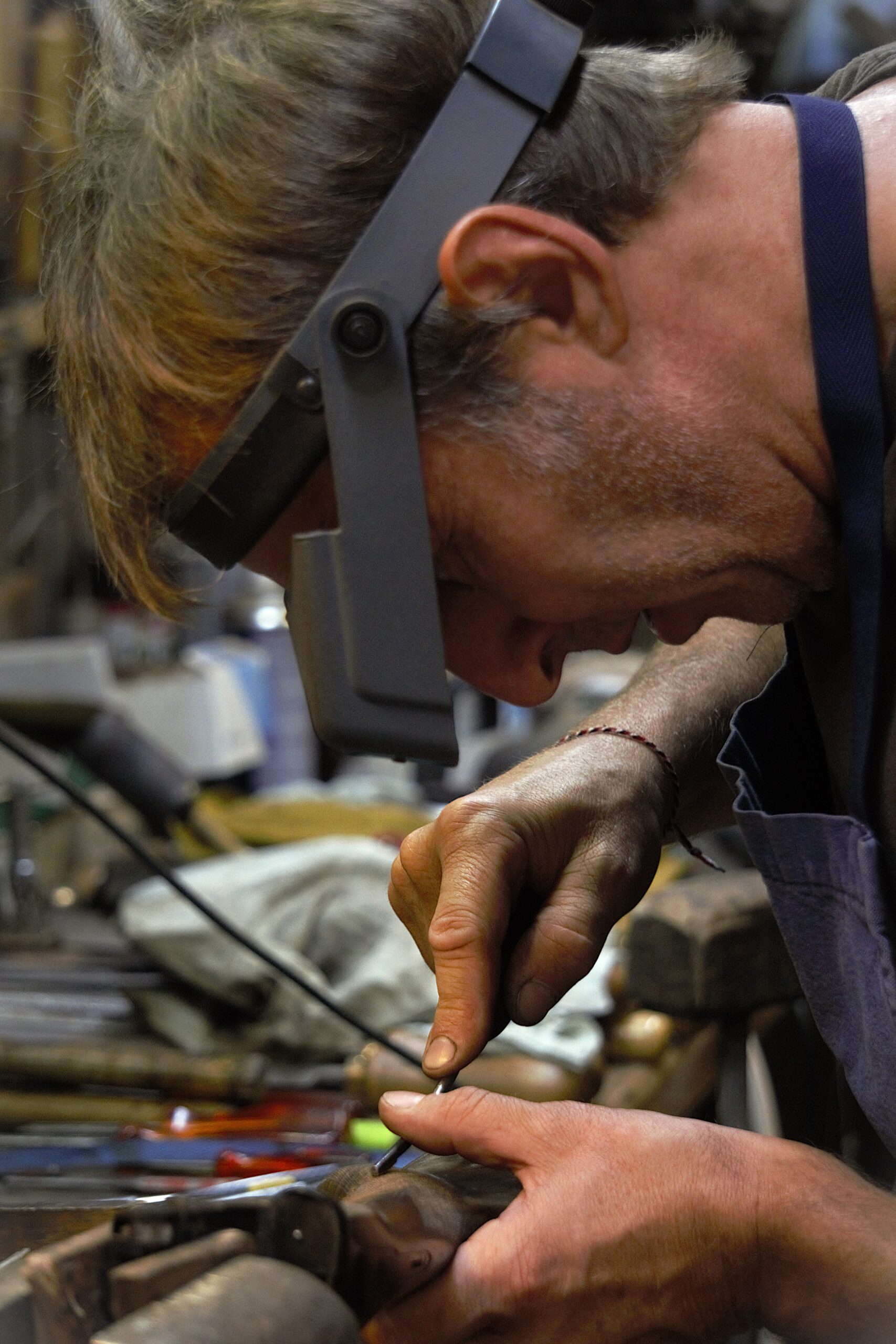 Artisan concentré portant une loupe frontale, ses mains réalisent un travail de précision avec un outil sur un objet dans un atelier.