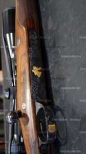 Carabine de chasse en bois précieux avec gravures. Récepteur noir orné d'une tête d'orignal dorée. Culasse et lunette de tir visibles.