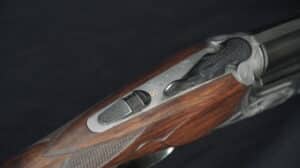 Détail de la crosse en bois et du récepteur métallique gravé d'un fusil de chasse, montrant le sélecteur de sécurité et un 'S' doré.