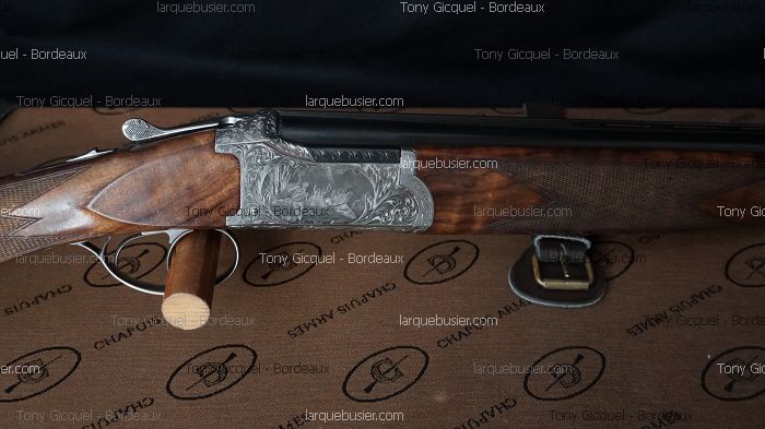Fusil de chasse double canon en bois et métal gravé, sur tapis marron Chapuis Armes avec bouclerie laiton.