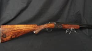 Fusil de chasse juxtaposé détaillé. Crosse en bois clair veiné, boîtier de culasse gravé de trois cercles, posé sur un support.