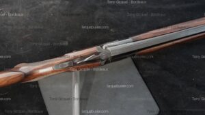 Fusil ou carabine double à bascule ouverte, crosse en bois finement travaillée et mécanisme métallique apparent sur fond sombre.