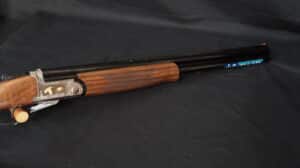 Fusil de chasse avec gravures dorées de canards sur le récepteur, crosse en bois quadrillée et canons noirs. Ruban "FAIR".