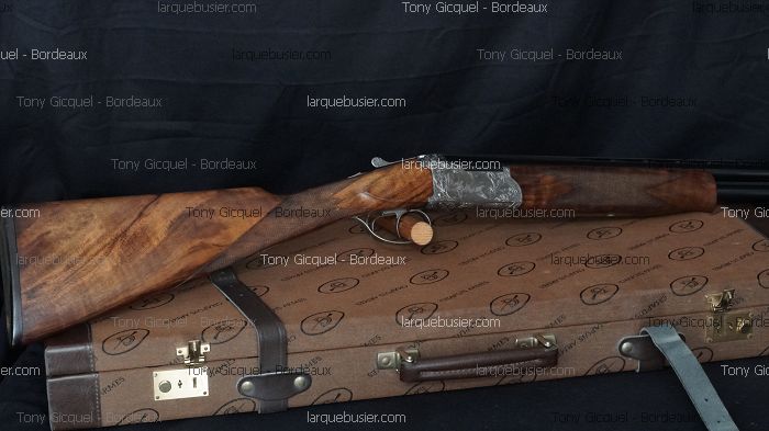 Fusil de chasse superposé, crosse et devant en bois, platines gravées, posé sur une mallette marron. Textes Tony Gicquel - Bordeaux, larquebusier.com.