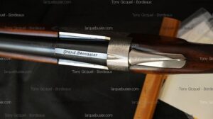 Gros plan détaillé sur la culasse et les canons brunis d'un fusil de chasse, avec l'inscription gravée 'Grand Bécassier' sur la bande supérieure métallique.