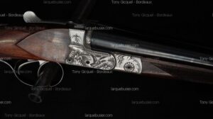 Fusil de chasse gravé : canard, motifs floraux sur platines, bois de crosse. Texte "MERLEDA", "Tony Gicquel - Bordeaux", "larquebusier.com".