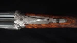 Fusil de chasse double canon, gravures florales argentées détaillées sur la culasse et bois de crosse figuré.