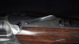 Détail du boîtier métallique d'un fusil de chasse, orné de gravures artistiques représentant des plumes et un motif stylisé 'S'. La crosse en bois poli est visible en dessous.