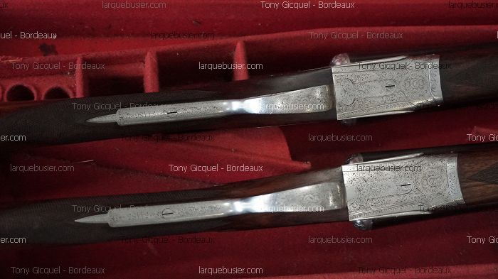 Deux actions de fusil de chasse gravées, avec bois foncé et métal ciselé, posées dans un étui doublé de velours rouge. Textes visibles: 'Tony Gicquel - Bordeaux', 'larquebusier.com'.
