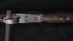 Récepteur et pontet d'un fusil de chasse gravé à l'anglaise, avec incrustations florales en argent et crosse en bois texturé. Texte visible: 'M'Me...'