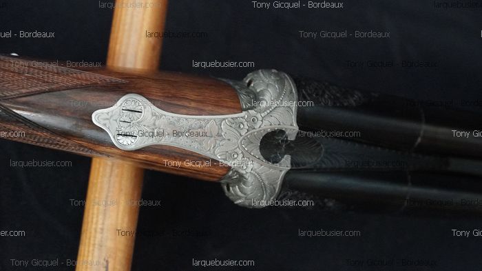 Détail d'un fusil de chasse: mécanisme en métal gravé de motifs floraux, crosse en bois veiné. Texte: Tony Gicquel - Bordeaux, larquebusier.com.