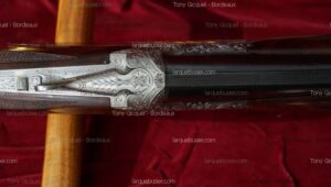 Mécanisme de fusil de chasse luxueux finement gravé (motifs floraux, argenté), crosse bois, canon texturé, sur velours rouge. Tony Gicquel - Bordeaux, larquebusier.com.