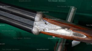 Gros plan sur un fusil de chasse double canon, crosse en bois et culasse en acier finement gravé. Mentions "Tony Gicquel - Bordeaux" et "larquebusier.com" sont visibles.