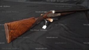 Fusil de chasse juxtaposé ancien à crosse en bois de noyer et platines gravées, posé sur tissu sombre, avec étiquette. Marque Tony Gicquel - Bordeaux, larquebusier.com.