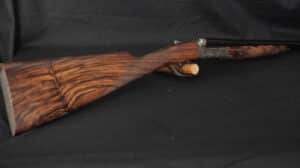 Élégant fusil de chasse juxtaposé sur fond noir. Crosse et devant en bois de noyer veiné, bascule en métal gravé.