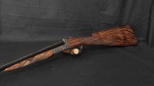 Fusil de chasse juxtaposé à double canon, crosse et devant en bois figuré, métal noir gravé, sur fond sombre plissé.