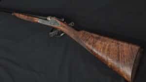 Fusil de chasse juxtaposé ancien avec canons noirs, platines gravées argentées et crosse en noyer figuré posé sur fond sombre.