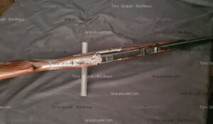 Fusil de chasse juxtaposé avec crosse en bois veiné et gravures sur la platine, posé sur tissu foncé. Vendu par Tony Gicquel - Bordeaux, larquebusier.com.