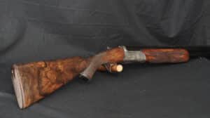 Fusil de chasse superposé, crosse en bois de ronce de noyer veinée, bascule argentée gravée, sur fond noir. Élégant et classique.