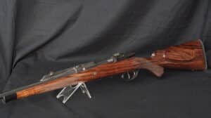 Fusil de chasse à crosse en bois figuré et canon en acier, posé sur un support transparent sur fond noir uni.