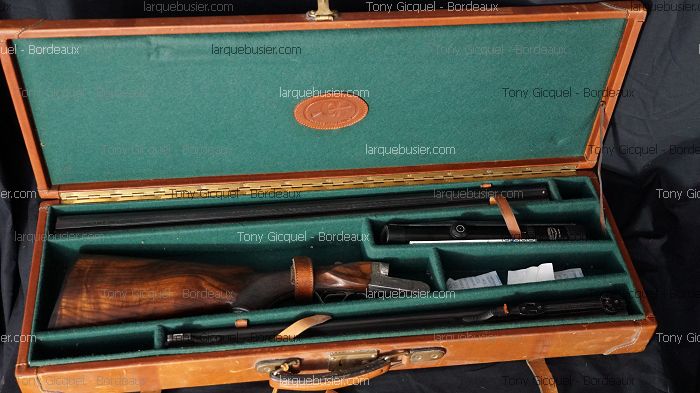 Fusil de chasse démonté dans une mallette en cuir marron avec intérieur en feutre vert. Crosse en bois, canons en acier, et logo en cuir sur le couvercle.