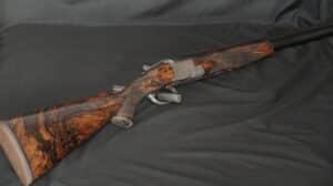 Fusil superposé haut de gamme en bois madré richement veiné et culasse métallique finement gravée, sur fond noir.