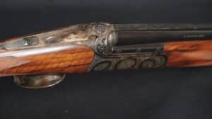 Fusil de chasse superposé, crosse bois et devanture ornées de gravures fines. Boîtier de culasse ciselé avec motifs détaillés.