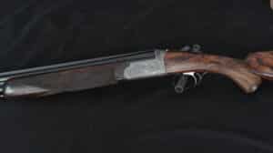 Fusil de chasse superposé avec bascule en argent richement gravée, crosse et devant en bois de noyer flammé quadrillé, sur fond noir.