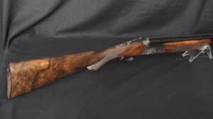 Fusil superposé de luxe avec crosse en bois de noyer veiné et récepteur métallique finement gravé, posé sur fond sombre.
