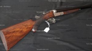 Fusil de chasse superposé, crosse bois brun, pontet métallique. Étiquette blanche. Receveur argenté, canons noirs, aspect classique.