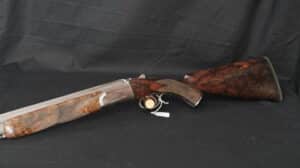 Fusil de chasse juxtaposé ou superposé avec crosse et devant en bois figuré sur fond sombre. Détails argentés et plaquette visible.