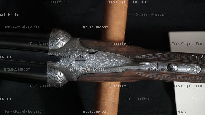 Gros plan sur la bascule ouvragée d'un fusil de chasse, avec gravures ornementales et crosse en bois quadrillée. Texte: Tony Gicquel - Bordeaux.