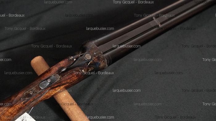 Fusil double-canon de chasse ancien, crosse en bois veiné et mécanisme en métal, posé sur un support en bois.